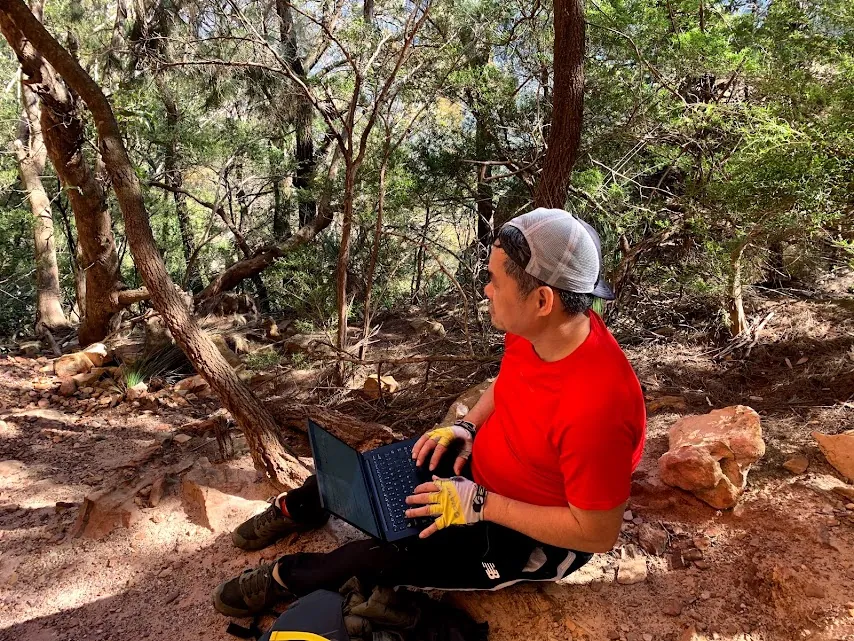 A hiker using a laptop outdoors beside a bush track
