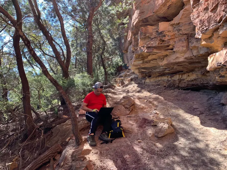 A hiker sitting on a rocky ledge beside a sandstone cliff, working on a laptop