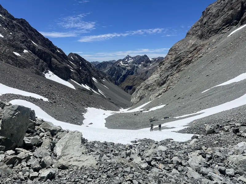 Two hikers crossing a snowfield in a steep rocky mountain pass, with alpine peaks beyond